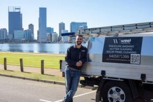 Perth-Pressure-Washing A man leans against a utility lorry labelled with cleaning services and contact details, with a city skyline and waterfront in the background.