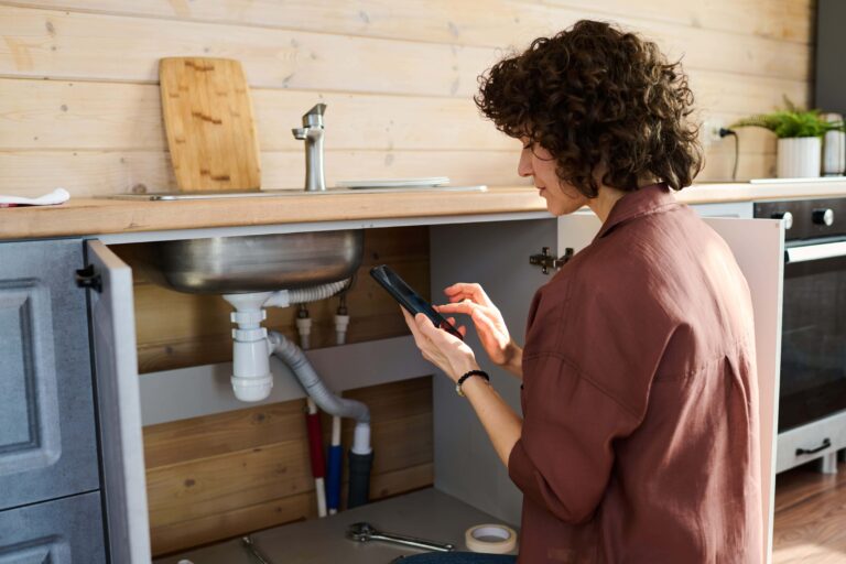 Young woman using a phone to report a plumbing issue under the kitchen sink-Backflow Prevention Burns Beach